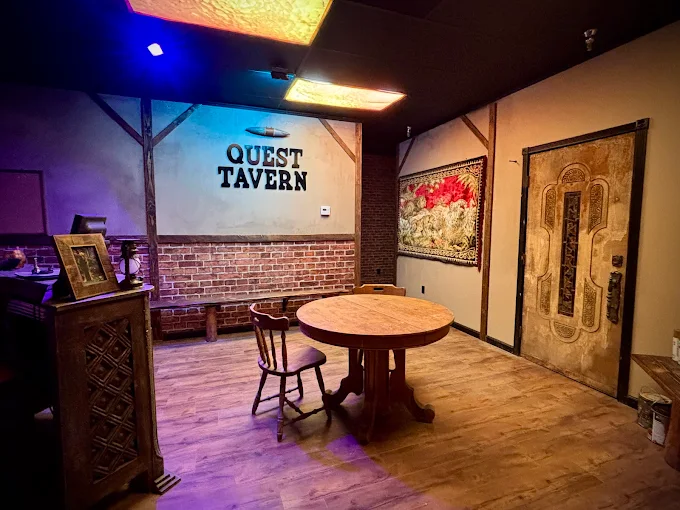 Quest Tavern Escape Room lobby in Upland, CA with round wooden table, chairs, brick wall, and Quest Tavern sign.